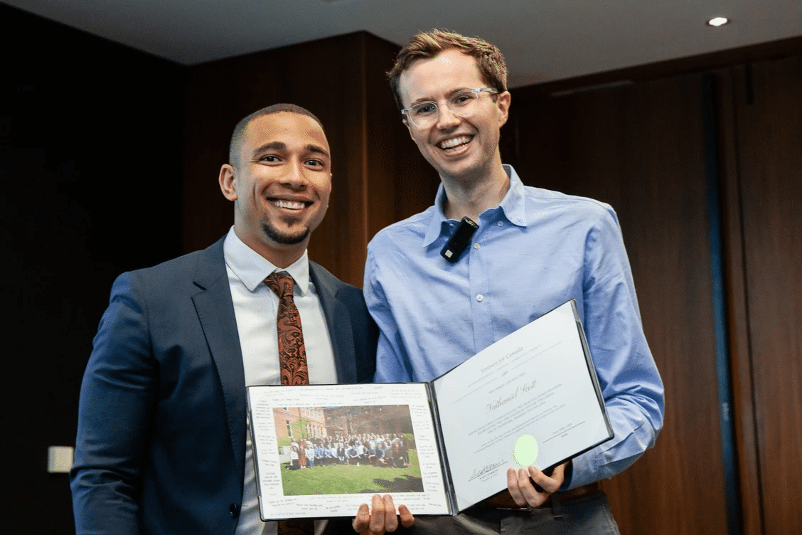 Ashoka Fellow Scott Stirrett (right, lighter skin glasses and shorter brown hair) with Nathaniel Scott (left, brown skin, dark short hair in a fade, goatee). Both people dressed in formal wear smiling at the camera. Background 