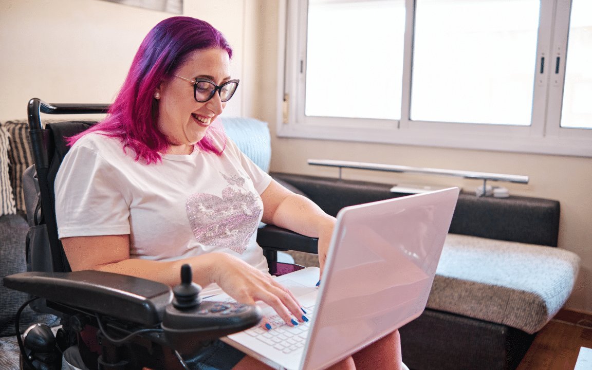 A person with pink hair uses a laptop while seated in a powered wheelchair in a bright living room.