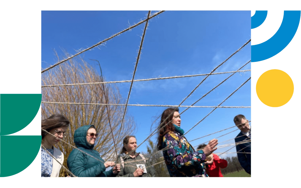 A group of people stands outdoors beneath a web of intersecting strings stretched between them. The activity takes place in an open, grassy area under a clear blue sky. The photo is framed by green, yellow, and blue geometric shape