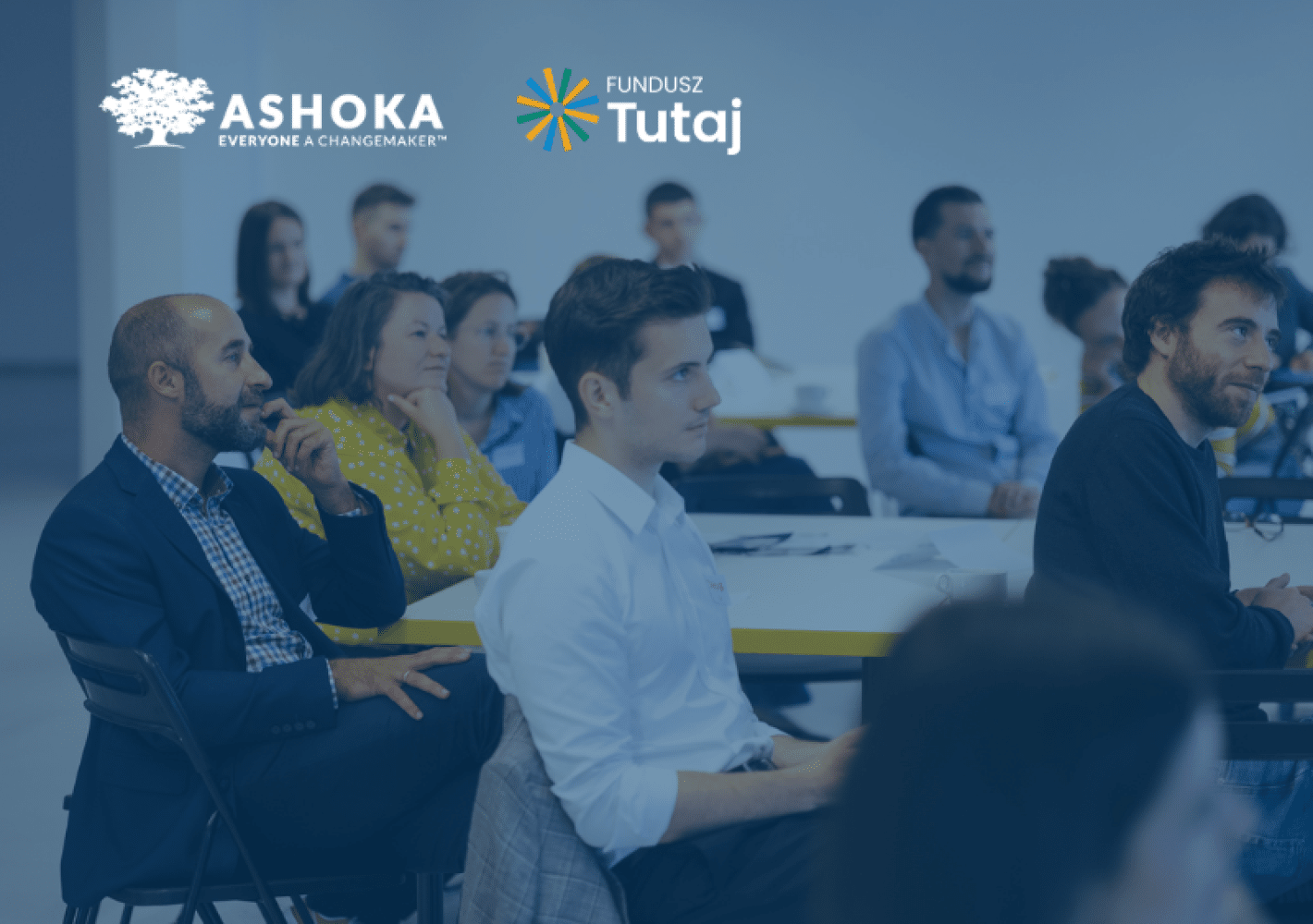 Zdjęcie przedstawia grupę osób siedzącą przy stole i słuchającą osoby, która jest poza kadrem. Na zdjęciu jest delikatny niebieski filtr. W lewym górnym rogu jest logo Ashoki z podpisem "Everyone a changemaker" i obok logo Funduszu Tutaj.