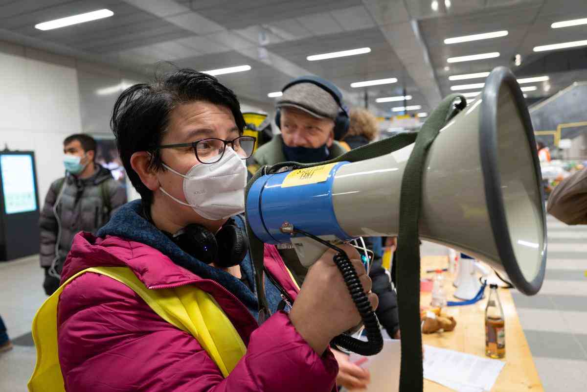 Person with short dark hair and glasses and a COVID mask speaking through a megaphone in an indoor space; background looks like either a big department store or a lobby in an office building