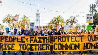 Photo of a faith and climate change protest. Large banner in front saying "People of Faith Rise Together to Protect Our Common Home." Lots of protest signs behind it with a number of people from all different faiths walking on a street. Background of palm trees, and a clock tower