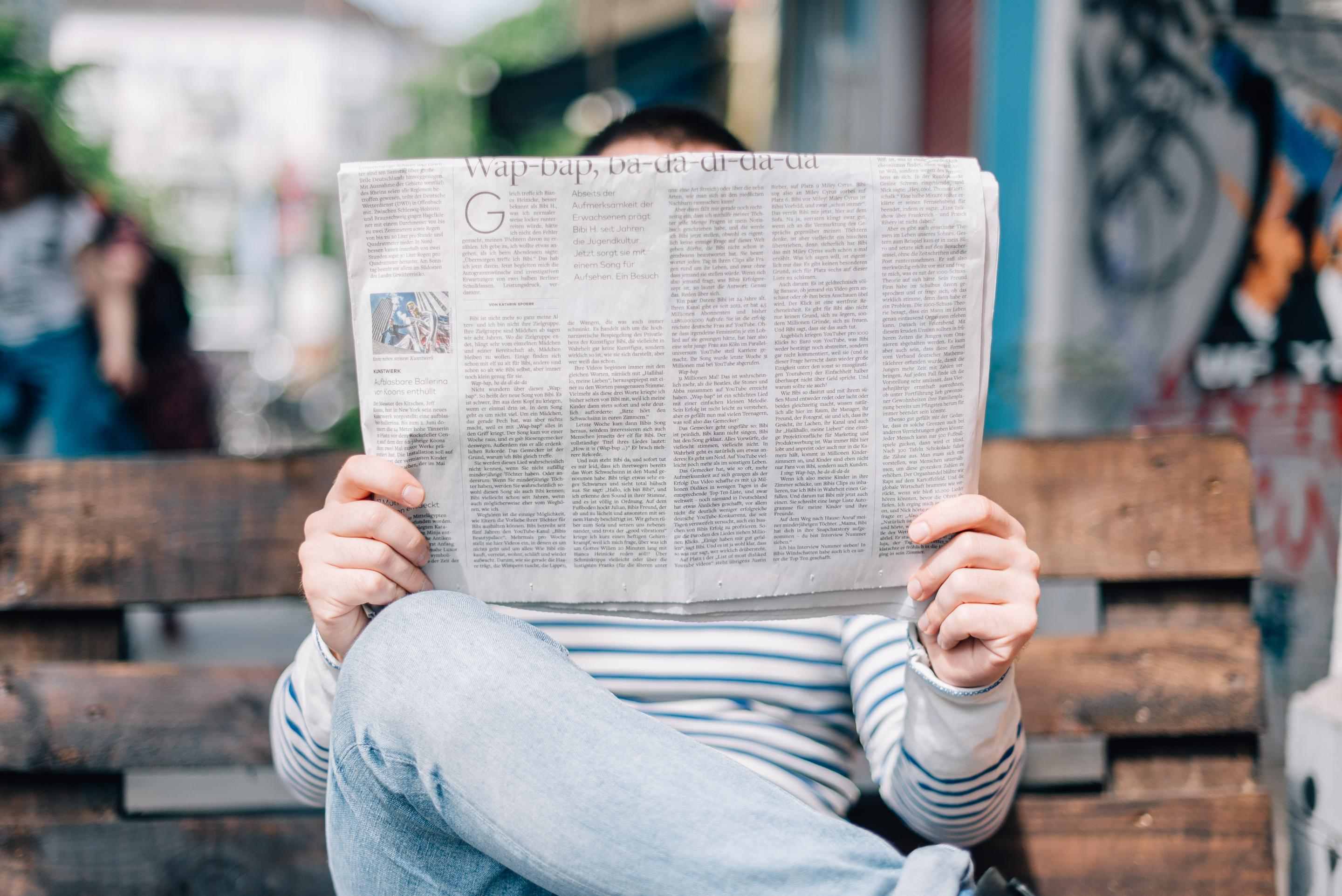 The photo shows a person sitting on a bench reading a newspaper. Their face is cover by the newspaper.