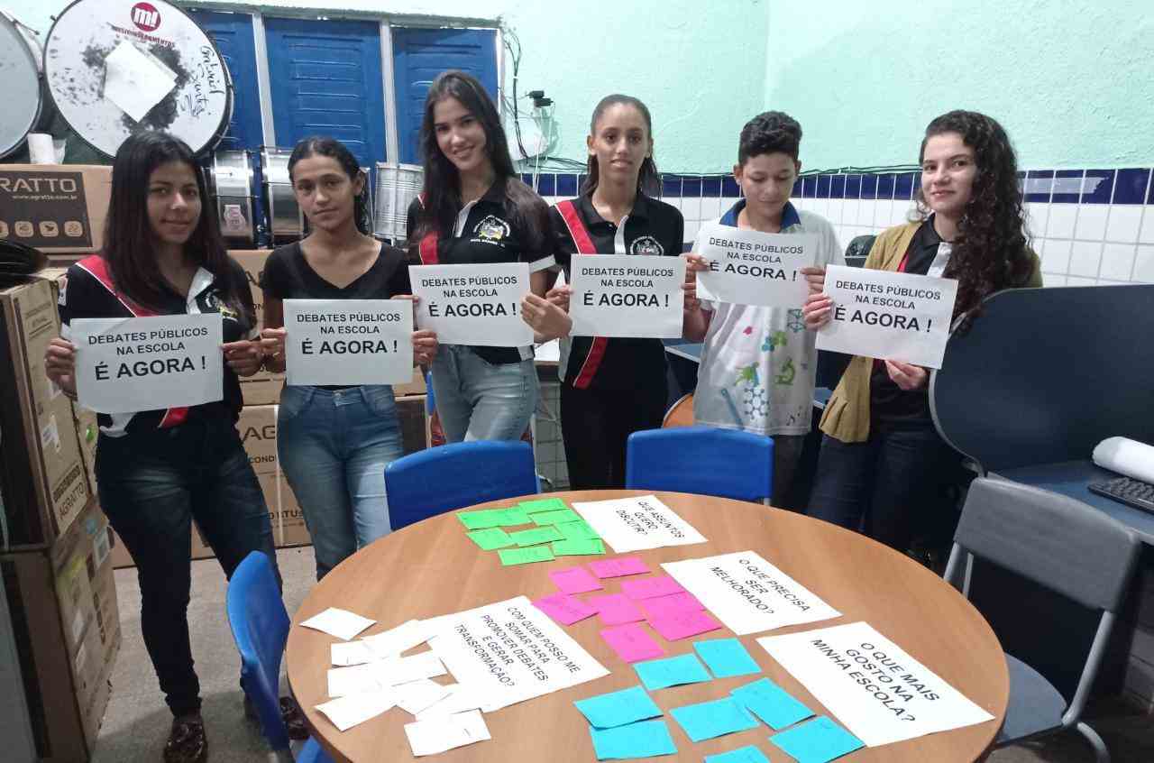 Grupo de jovens seguram cartazes sobre cultura democrática. À sua frente, há uma mesa com diversas folhas de papel com notas adesivas