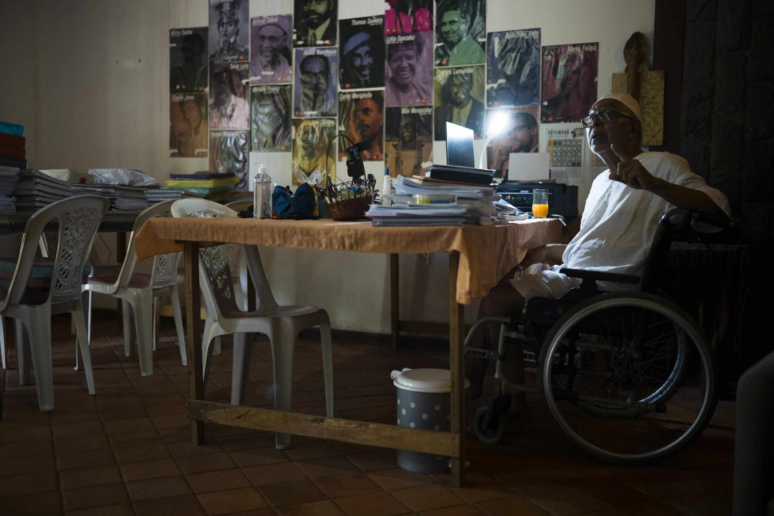 Person in a wheelchair speaking to someone to the camera's left. Backdrop of a desk with a wall covered in photos