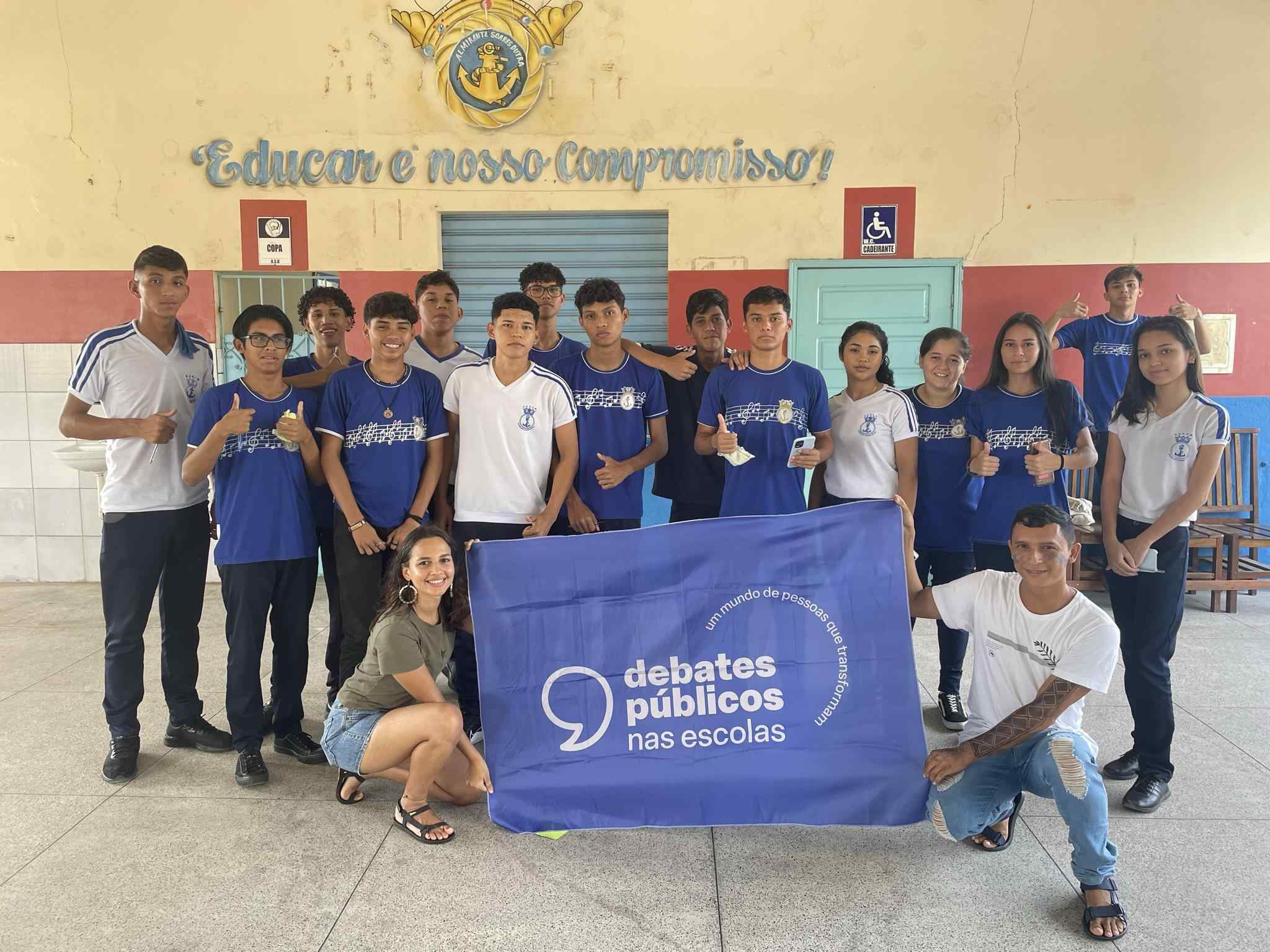 Grupo de jovens reunidos em um pátio. A turma sorri para a foto e mostra a bandeira dos Debates Públicos nas Escolas