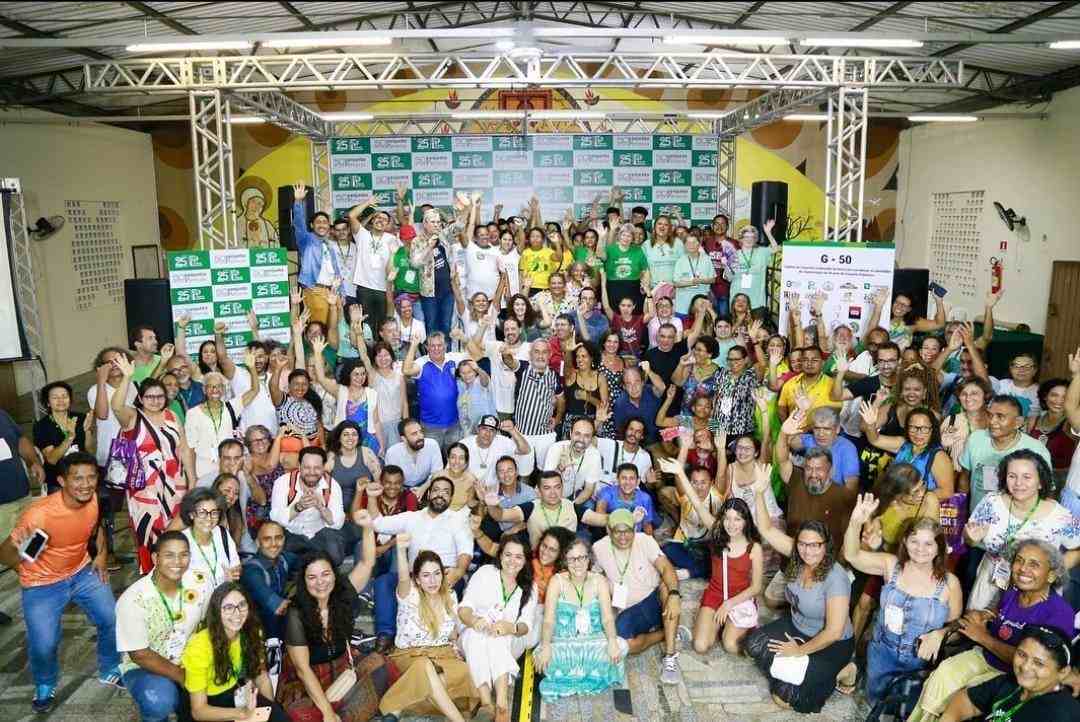 Grande grupo de pessoas reunidas em um salão. Ao fundo há um palco com uma estrutura de metal e um painel com retângulos verdes e brancos