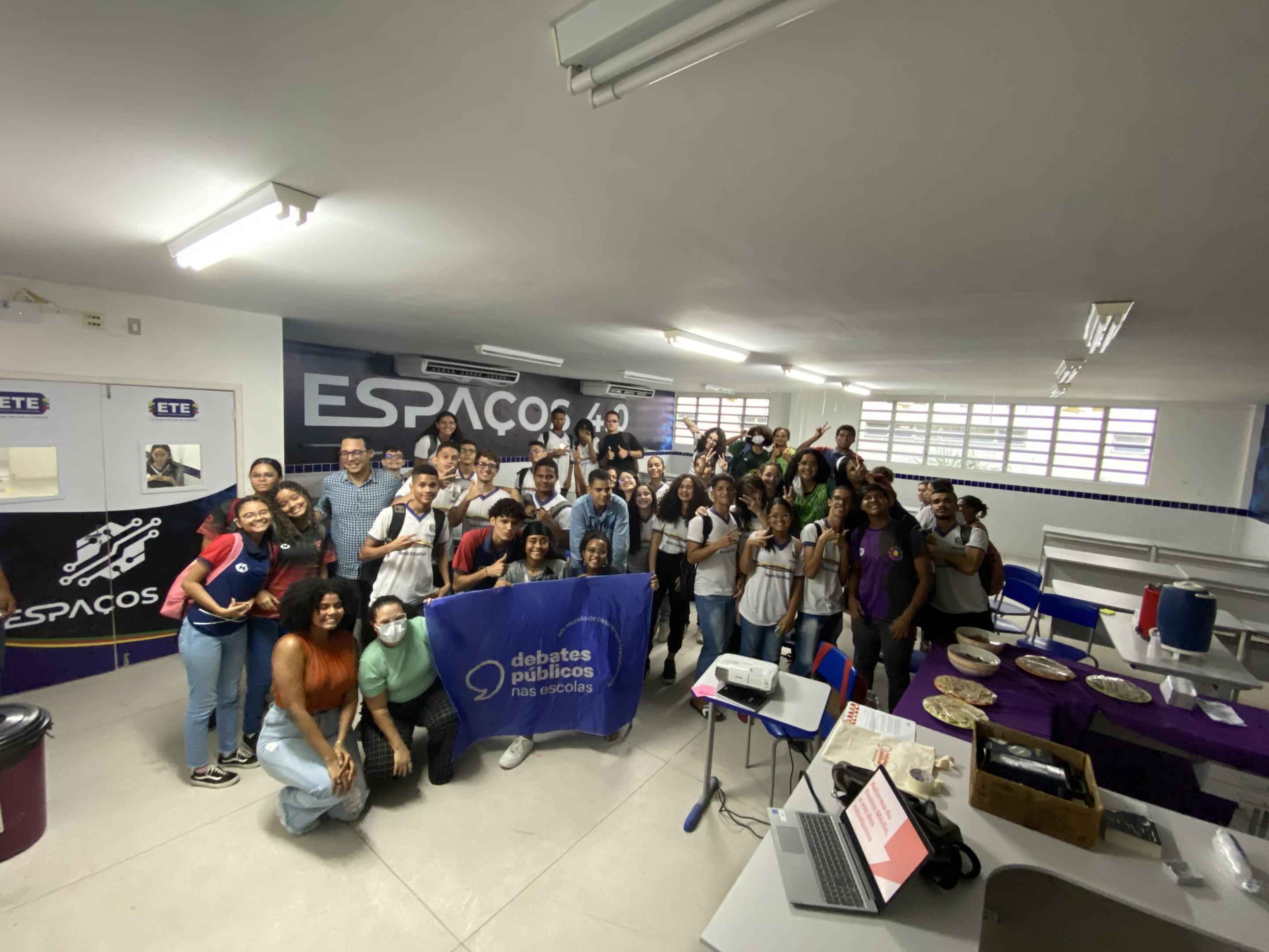 Grupo de jovens reunidos em uma sala de aula. A turma sorri para a foto e segura uma bandeira dos Debates Públicos nas Esocolas