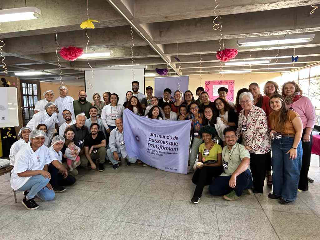 Grupo com cerca de 40 pessoas reunidas em uma ampla sala. As pessoas que estão no centro seguram a bandeira do Território Transformador de São Paulo