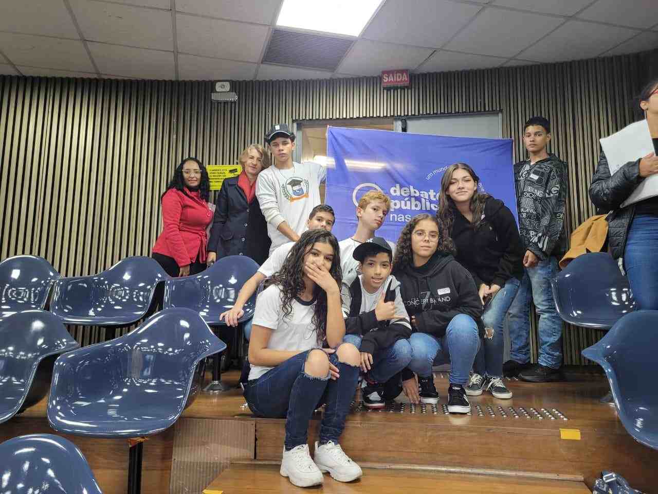 Grupo de jovens posa para a foto em um auditório. Ao fundo, dois deles seguram a bandeira dos Debates Públicos nas Escolas