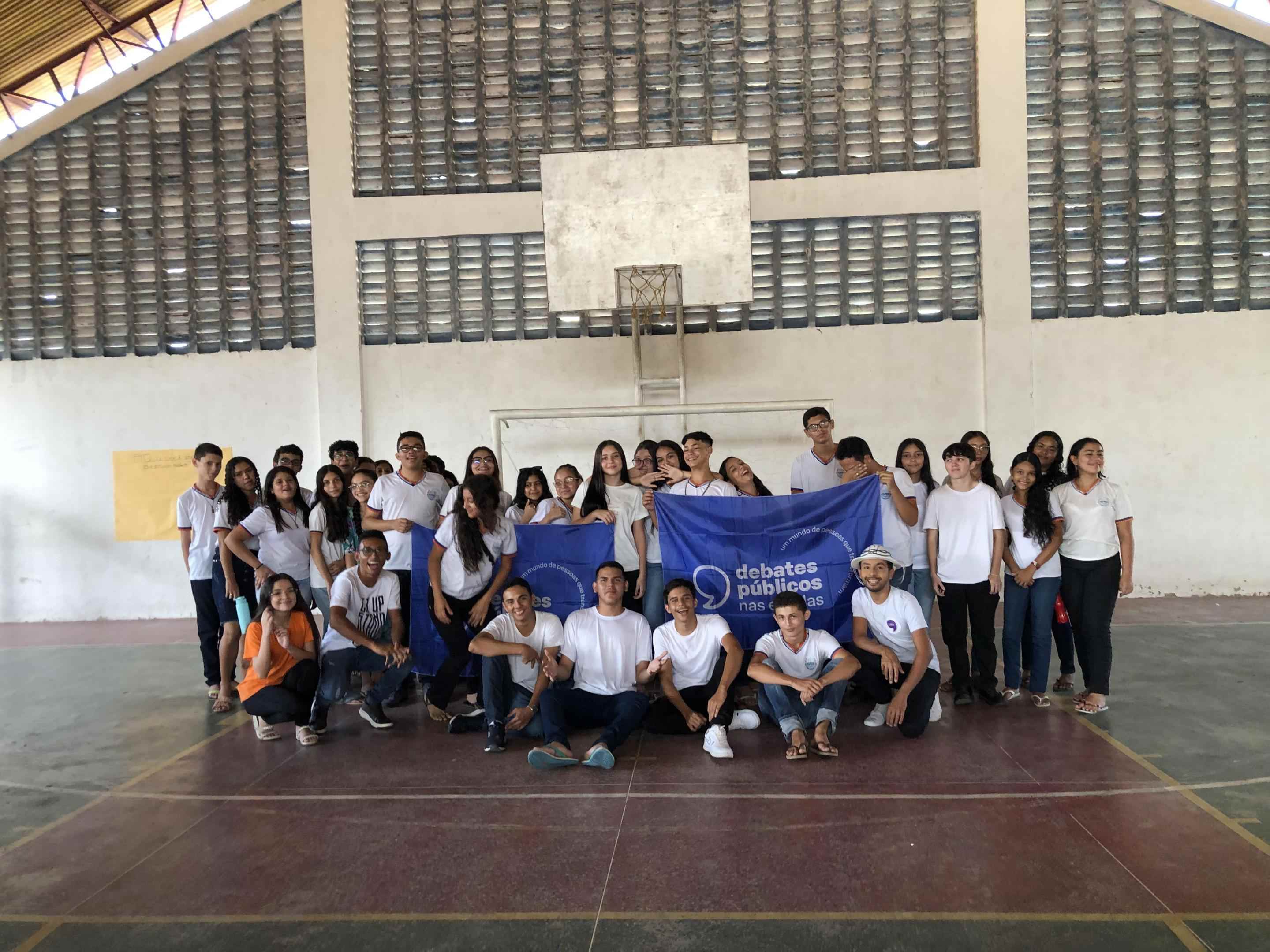 Grupo de jovens reunidos em uma quadra de escola. O grupo segura a bandeira dos Debates Públicos nas Escolas