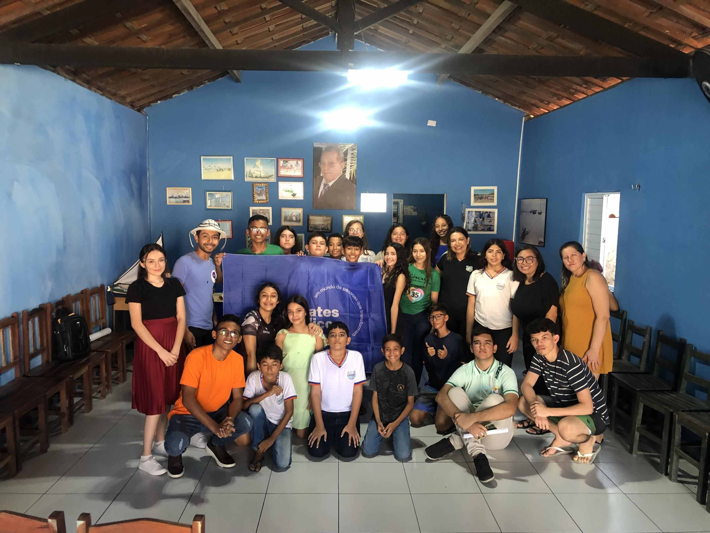 Grupo de jovens reunidos em uma sala de aula. A turma sorri para a foto e mostra a bandeira dos Debates Públicos nas Escolas