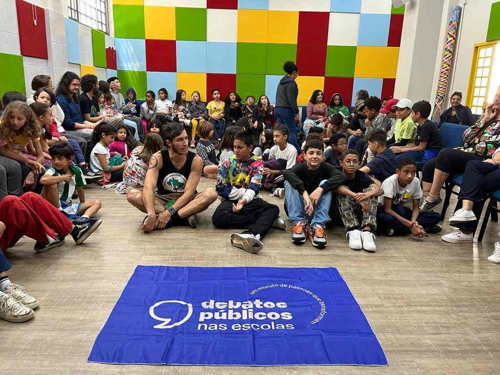 Grupo de crianças e adultos reunidos em uma sala com paredes coloridas. No chão, está a bandeira dos Debates Públicos nas Escolas