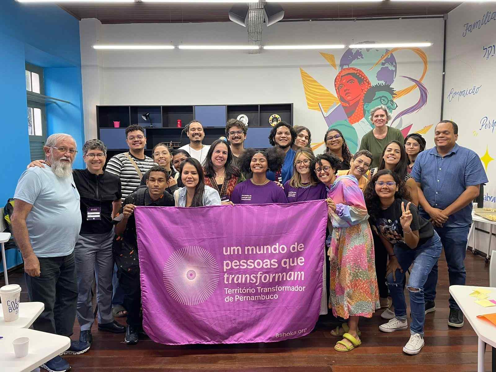 Grupo de pessoas reunidas em uma sala. Elas sorriem para a foto e seguram a bandeira do Território Transformador de Pernambuco
