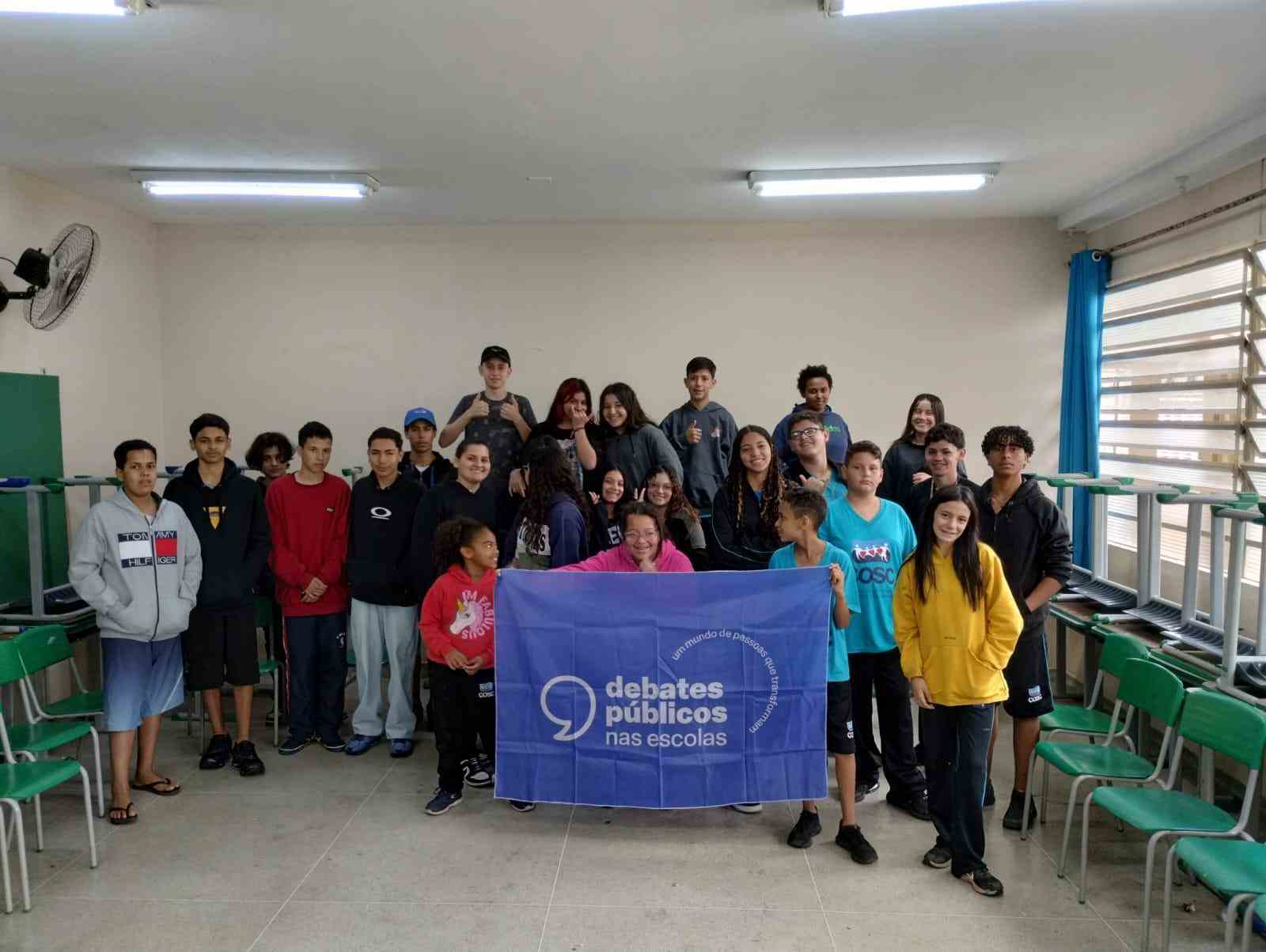 Grupo de jovens reunidos em uma sala de aula. A turma sorri para a foto e mostra a bandeira dos Debates Públicos nas Escolas