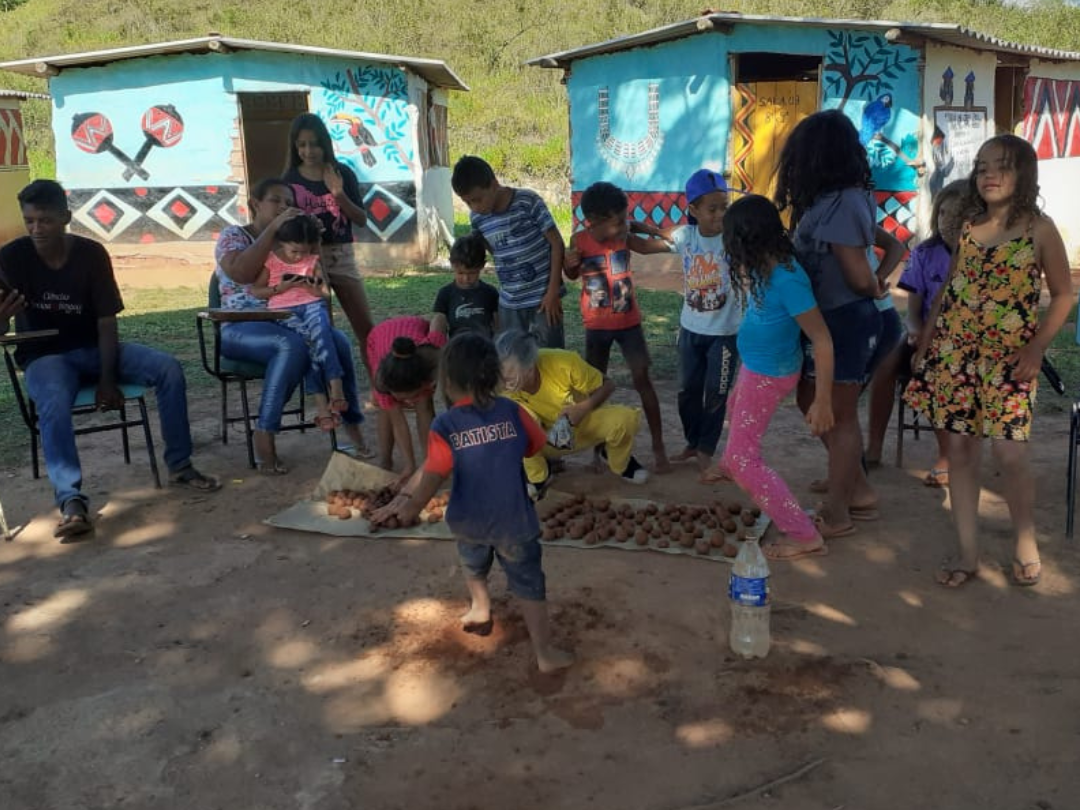 Crianças e adultos reunidos ao ar livre. Algumas crianças brincam na terra e algumas pessoas estão sentadas em cadeiras. Ao fundo, há pequenas casas decoradas com grafismos indígenas