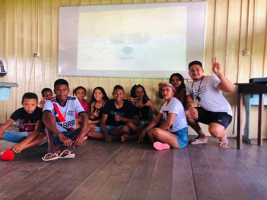 Crianças e jovens sentados no chão de uma sala de aula. O grupo sorri para a foto e ao fundo há um telão