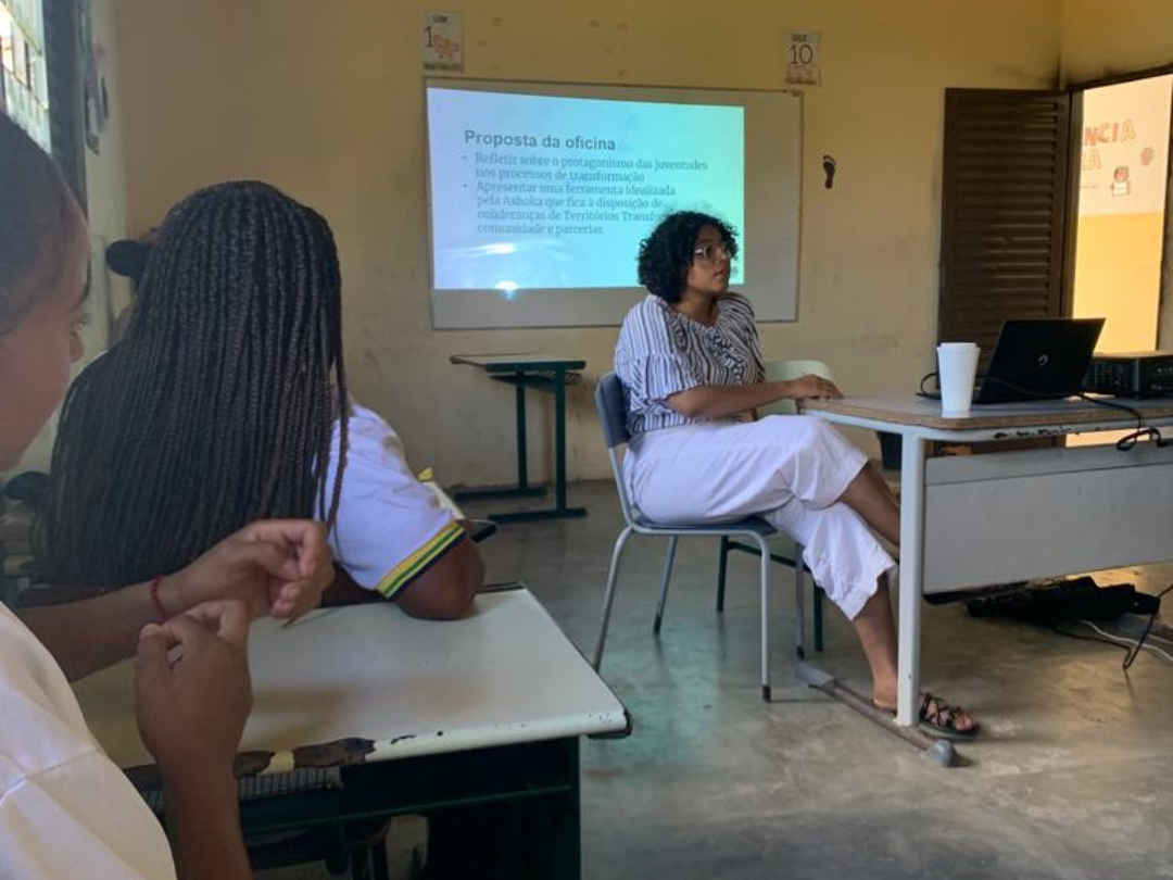 Ao centro, uma jovem negra de cabelos cacheados está sentada numa cadeira. À esquerda, duas jovens estão sentadas, de costas para a câmera, olhando para a outra jovem. Ao fundo, há um telão com uma projeção