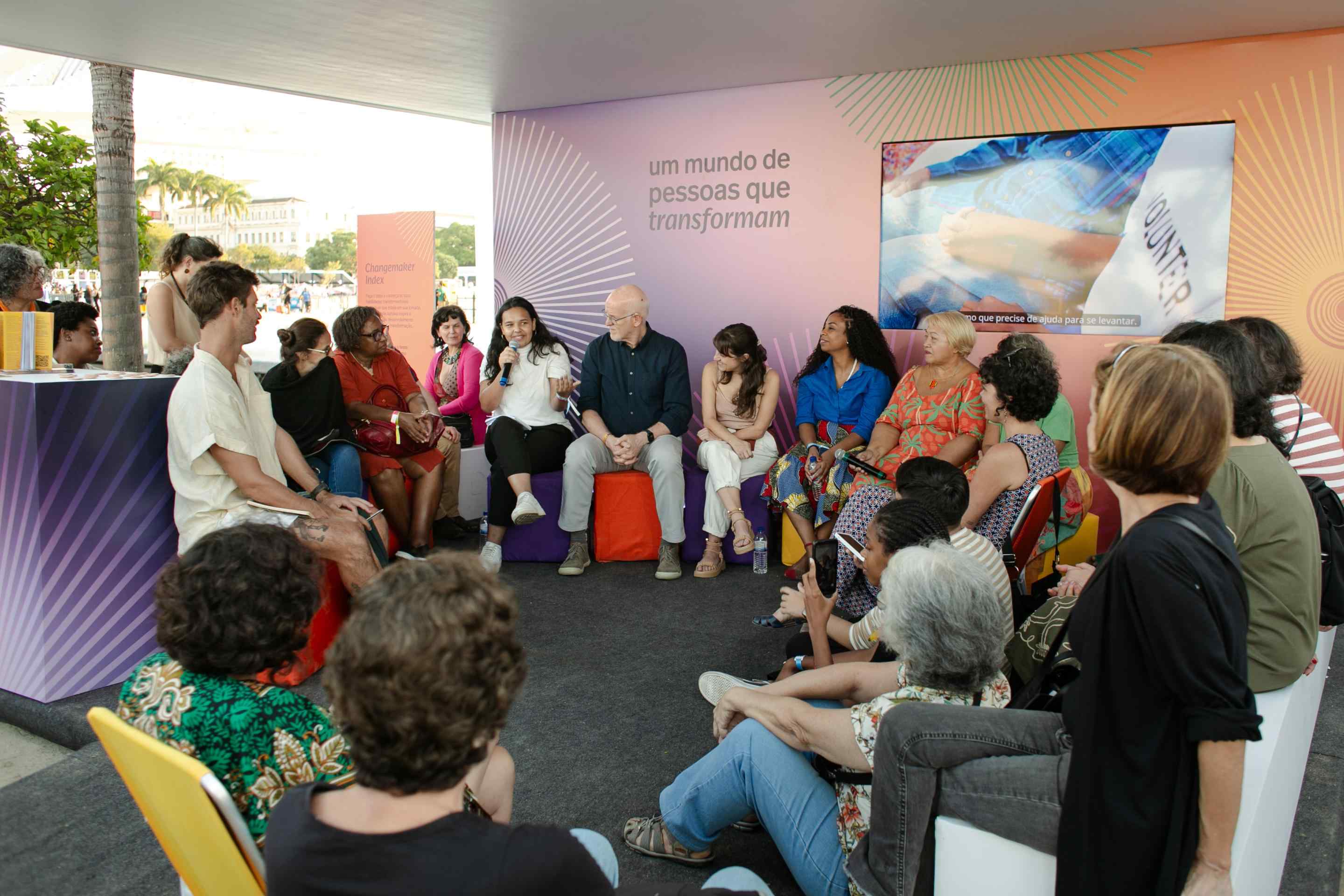 Grupo de pessoas reunida em um estande ao ar livre. Elas estão sentadas em roda, em pufes, bancos e cadeiras de praia. Ao fundo, há um painel colorido onde se lê "um mundo de pessoas que transformam" com uma televisão ao lado