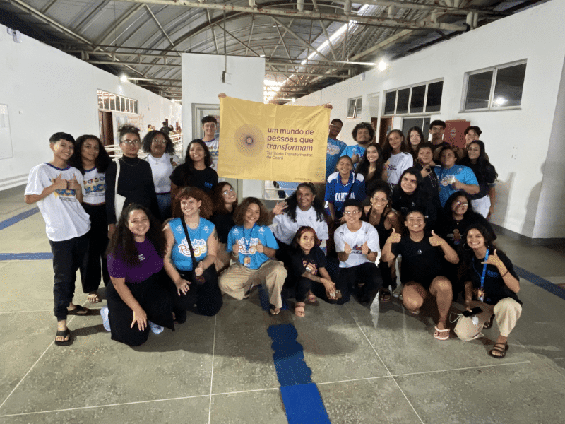 Grupo de jovens diversos reunidos em uma sala ampla. Ao fundo, duas pessoas seguram a bandeira do Território Transformador do Ceará