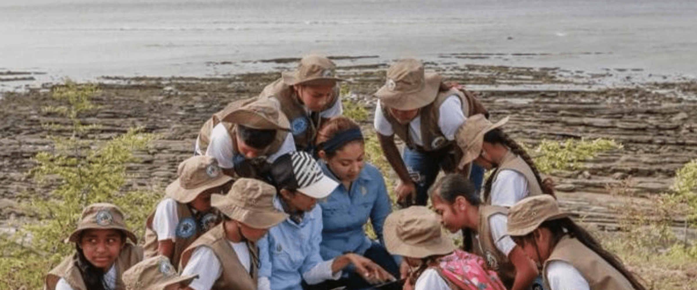 Una mujer que enseá a un grupo de niñas, niños y jóvenes. Se ve un espacio natural con lo que parece un lago y montañas de fondo. 