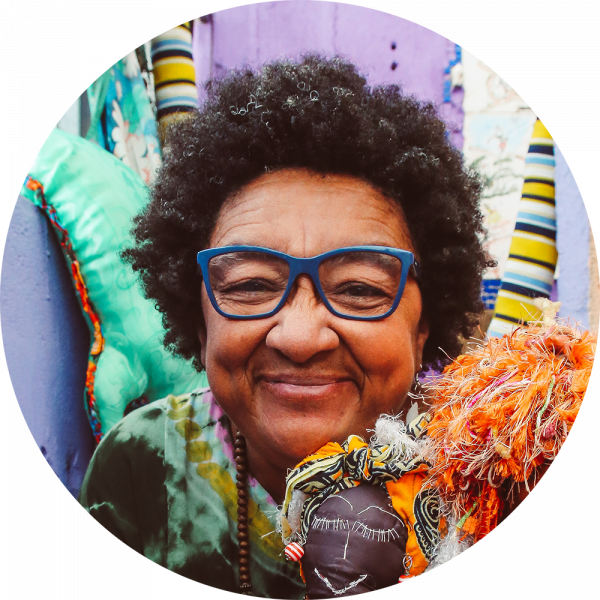 Mulher de óculos azuis, cabelo crespo, com leve sorriso, segurando uma boneca colorida.