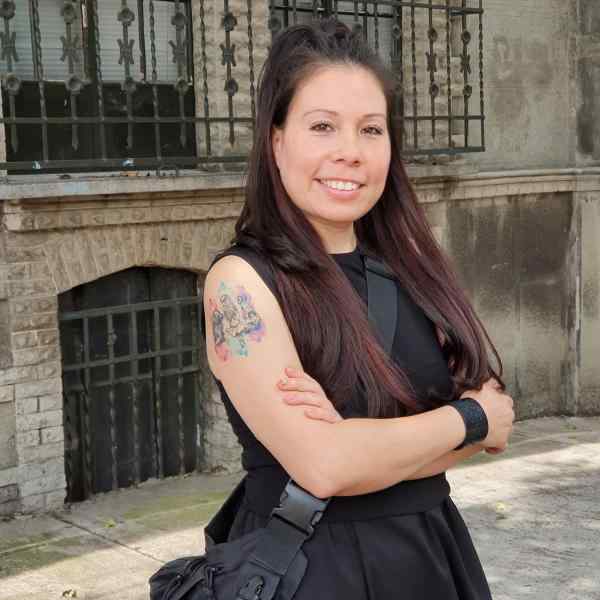 Photo of Iliana Luna, Ashoka Mexico Staff. Person with long dark hair smiling at camera, dressed in black dress with a black flowy satchel bag. Located in the middle of a sidewalk. In the background is the outside of a buidling, with bars on the basement door and windows, and outside walls are light beige