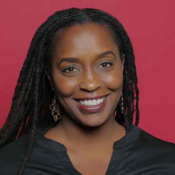 Photo of Angelou Eweilo, Ashoka Leadership Group Member for Empathy. Headshot of a person with dark skin and dark shoulder length hair smiling at a camera, dressed in a black blouse. Background is a red wall