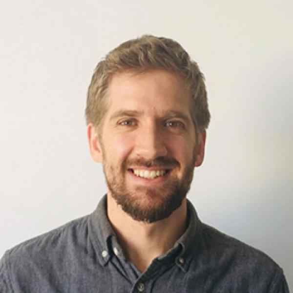 White man, with blonde hair and beard, wearing a dark gray shirt. He is smiling and the backroung is lighter gray.