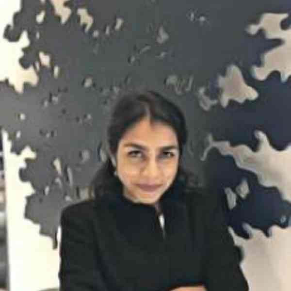 Woman with dark hair, wearing a black coat. In her background there's a white wall with Ashoka's tree in it. 