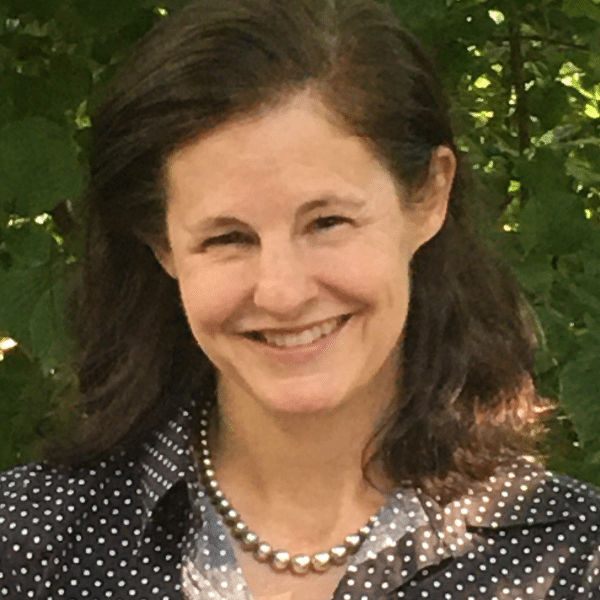 Photo of Emily Lawson, Leadership Group Member of Ashoka. Person with shoulder length brown hair and lighter skin smiling at the camera. Dressed in polka dot blouse; background of tree leaves