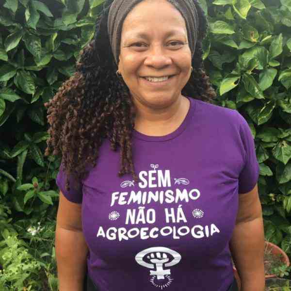 Beth is smiling, she has long curly dark hair and wears a purple T-shirt that reads 'Without feminism, there is no agroecology'