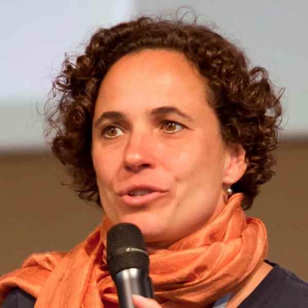 Photo of President Emerita Diana Wells; person with a bright orange scarft, short curly hair speaking on a panel; microphone held up in from of their chin.