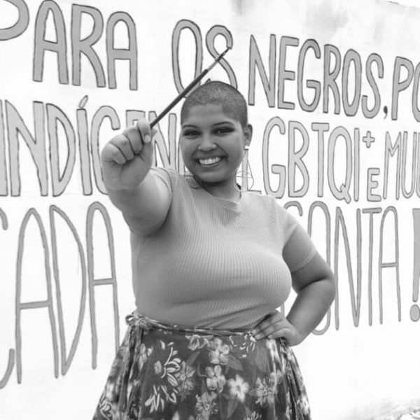 Beatriz is a black girl with shaved hair. She is wearing a T-shirt and a flowery skirt and she is holding a brush. Behind her there is a mural painted by her. The photo is black and white