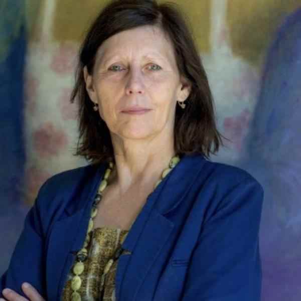 Portrait of Fellow Ingrid De Jonghe. She is wearing a long sleeve dark blue blazer jacket with a yellow shirt underneath. Her shoulder lenght straight dark brown hair is styled loose and she is smiling at the camera. The background is an out of focus mural painting .