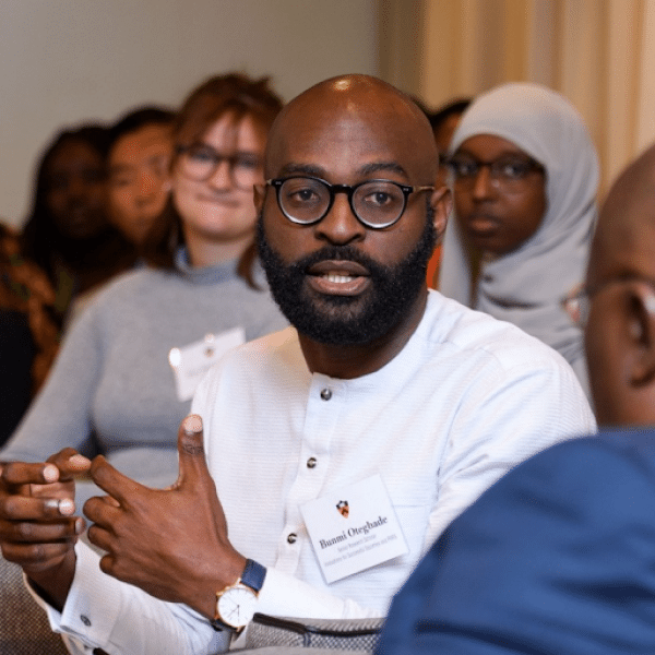 Headshot of Bunmi Otegbade. Darker skinned person with shaved head and beard talking to someone in the foreground