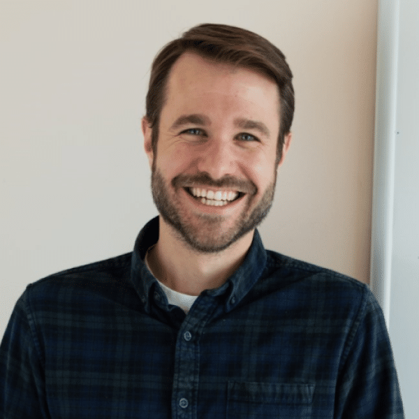 Headshot of Nick Boedicker. Person with lighter skin short hair and beard smiling at the camera. Dressed in a dark shirt, background of a wall