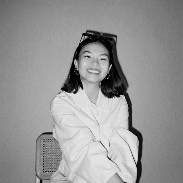 Photo of Yuxian Seow, Ashoka staff in the UK. Black and white photo of a person with shoulder length dark hair seated and smiling at the camera dressed in a light top and darker pants. Background of a wall