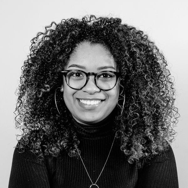 Foto em preto e branco de Gabriela, mulher negra de cabelos cacheados na altura do ombro. Ela sorri e usa óculos, blusa preta de gola alta e um colar