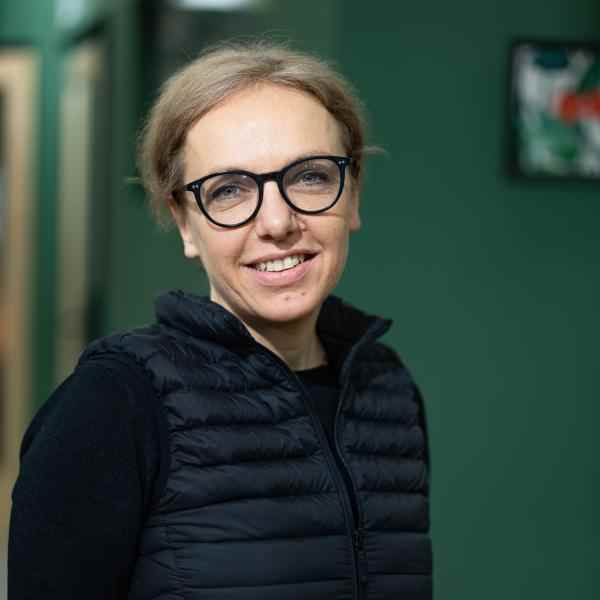 Zdjęcie przedstawia Marię Deskur. Kobieta ma ciemne okulary, blond włosy związane z tyłu głowy, czarną koszulkę z długim rękawem i czarny bezrękawnik. Uśmiecha się.