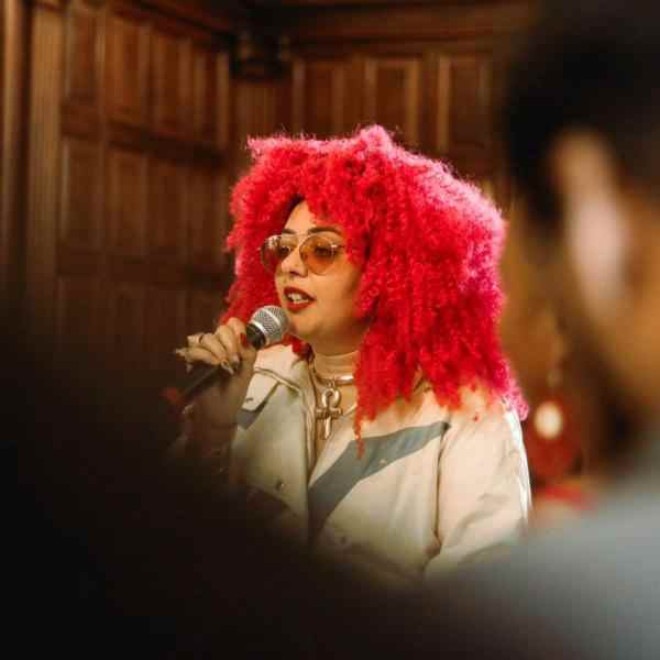 Black woman with big pink curly hair. She wears prescription glasses with dark, diamond-shaped lenses, red lipstick, a cream blouse with a high collar and a white jacket with gray details. She is standing in profile and holding a microphone to her mouth. 