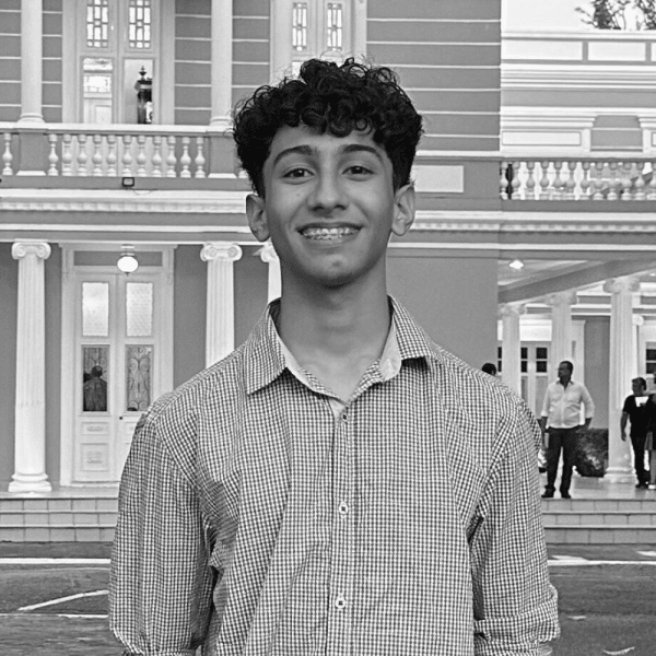 Um jovem com cabelo curto e com cachos, sorrindo para a foto, usando uma camisa social xadrez