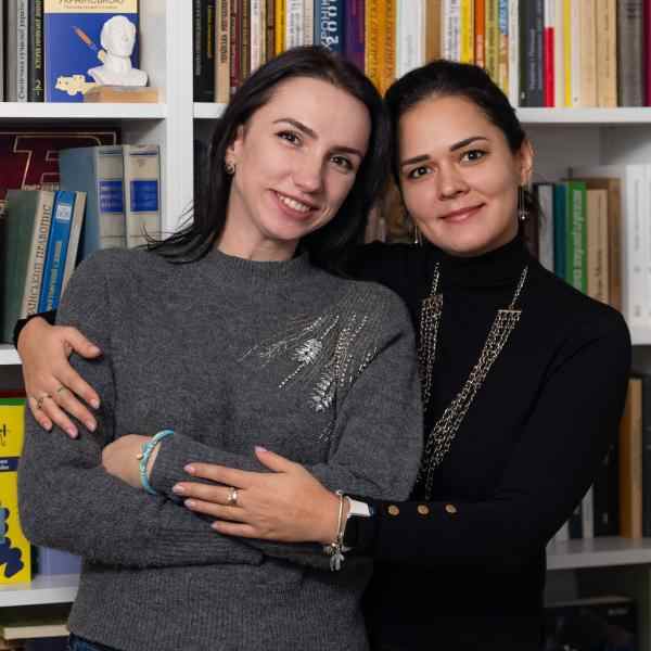Zdjęcie przedstawia Mariię Dehtiarenko i Tetianę Lysak, które obejmują się na tle regału z książkami.