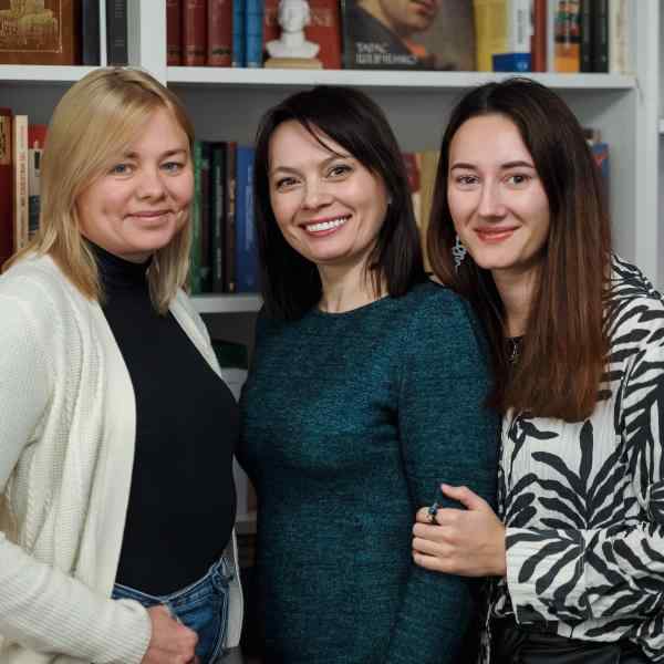 Zdjęcie przedstawia Tetianę Pakhaliuk, Nataliię Pokrovską, Varvarę Akhremenkę, które obejmują się na tle regału z książkami