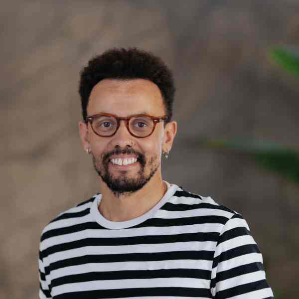 A photo of a black man with short curly hair and a neatly trimmed beard and moustache. He is wearing a striped long-sleeved T-shirt. He has a hoop piercing in his nose, hoop earrings, is wearing prescription glasses and is smiling for the photo. The background, which is a wall in earthy tones and apparently has a plant on it, is out of focus. 