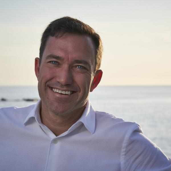 Photo of Jason Bernhardt-Lanier. Person with short dark hair smiling at the camera, dressed in a white top. background is an ocean and skyline