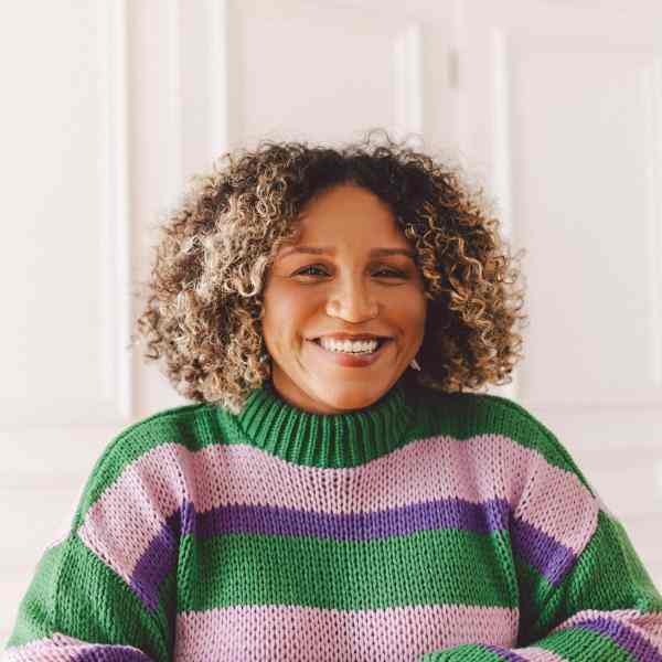 A photo of a black woman with short curly hair and blonde highlights. She is smiling and wearing a green and pink striped blouse. In the background, we see a white wall.