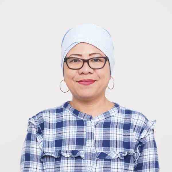 Person with white head covering and blue flannel shirt and jeans smiling at the camera. Seated on a white chair with a white background