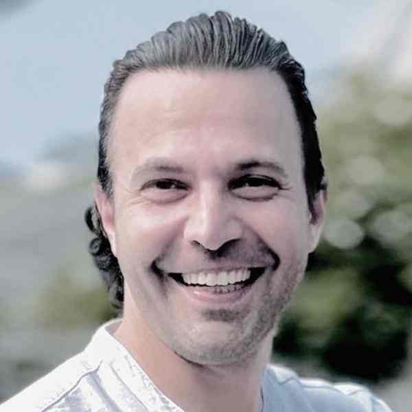 Profile photo of Ashoka Framework Change Staff Edward Edilbi. Photo of person with long slick backed hair smiling at the camera; background is of blurred out trees