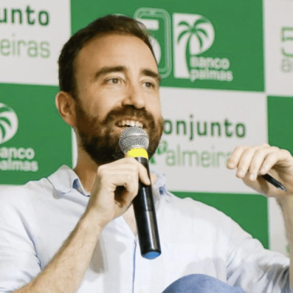 Man with a beard holding a microphone, smiling while speaking at an event. A green and white Banco Palmas and Conjunto Palmeiras-branded backdrop is behind him.